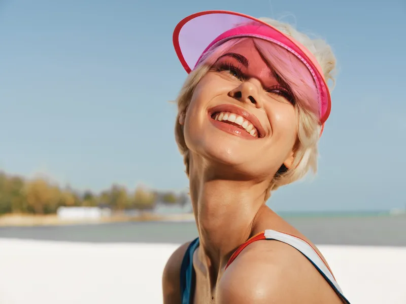 Bright portrait of a smiling woman on a sunny beach, wearing a pink visor. Warm light and blue sky create a carefree mood and vibrant summer atmosphere.