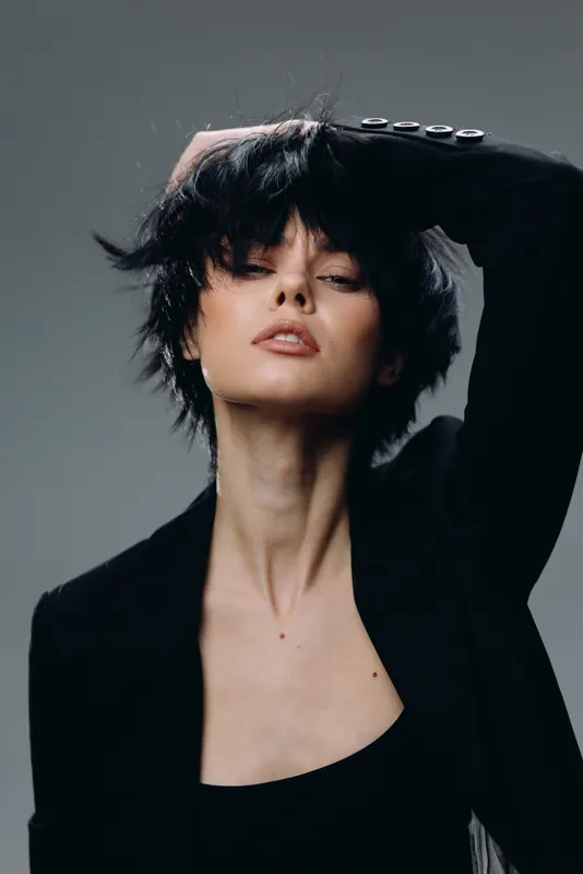 Stylish woman with short black hair wearing a black blazer posing confidently with hand in hair, expressing elegance and modern fashion.