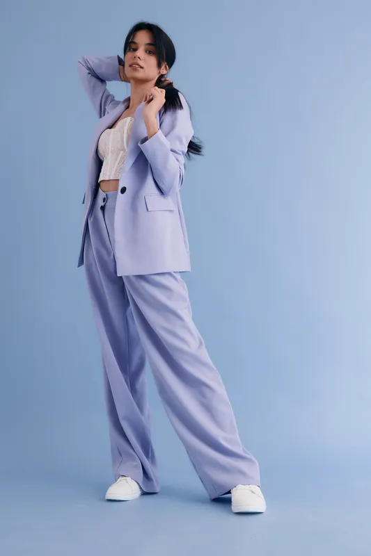 stylish woman in lilac suit posing confidently with hands behind head against plain blue background, casual white sneakers, modern fashion outfit