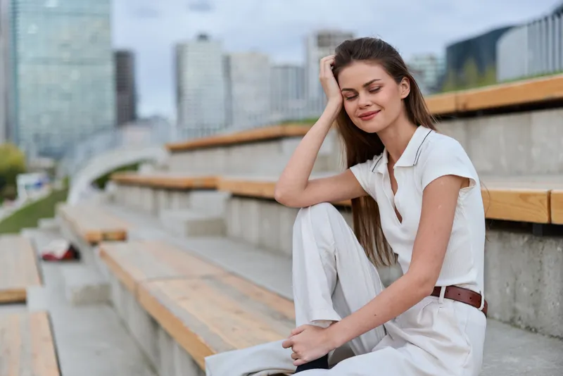 Young woman sitting relaxed outdoors in casual white outfit with modern urban background, smiling peacefully, lifestyle and fashion concept