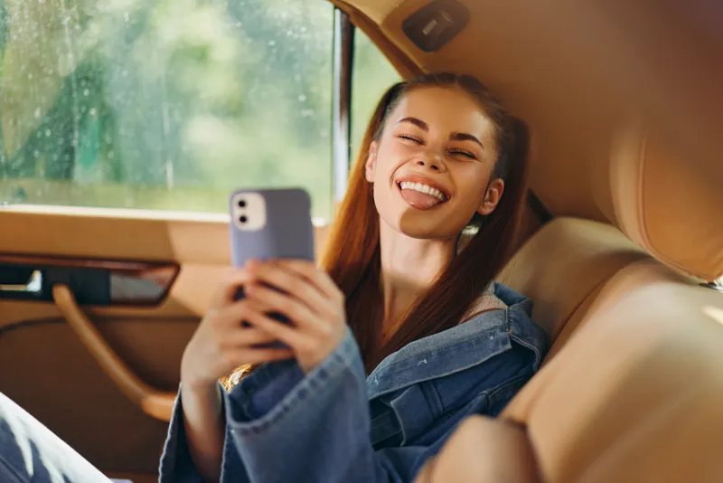 Happy young woman taking a selfie in denim jacket, sitting inside a car with a bright background, showcasing joy and confidence on a sunny day.