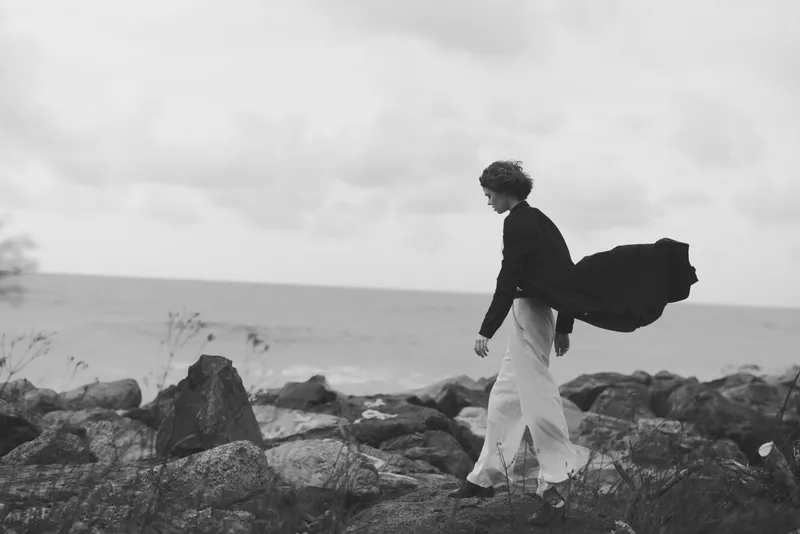 Woman walking along a rocky coast in a flowing cape, monochrome scene, wind twisting fabric, solitary mood, coastal landscape and fashion inspired movement, evoking quiet cinema aesthetics, dramatic