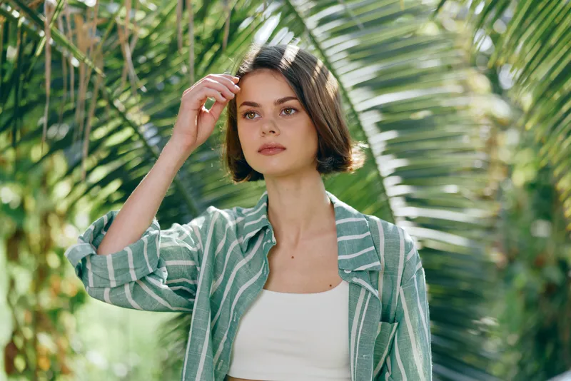 A woman outdoors among lush greenery, stylish striped jacket and white top, confident gaze, calm demeanor, fashion portrait capturing natural light and serenity.