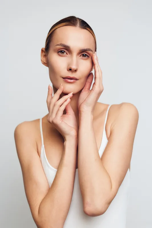 Young woman with clear skin posing with hands near face, calm expression, minimal makeup, wearing white tank top, studio lighting, neutral background, beauty and skincare concept