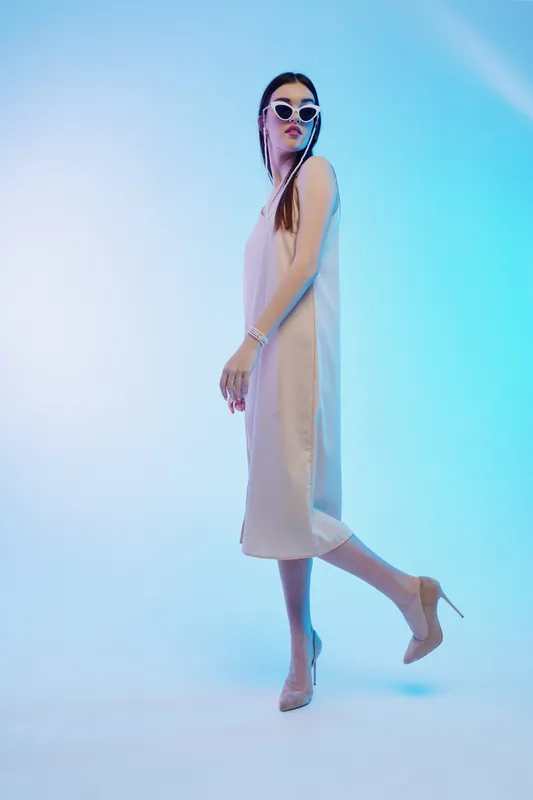 Stylish woman in a beige dress and sunglasses poses with one leg raised against a blue gradient background. Concept of woman beauty fashion and elegance.