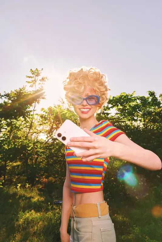 Smiling woman taking an outdoors selfie in bright sunlight, wearing striped crop top and light jeans. Retro fashion vibe, sunny mood, casual lifestyle, outdoor photography.