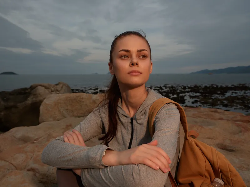 Young woman in a gray sports outfit reflecting on nature by the sea during sunset emoting serenity and determination, ideal for fitness and outdoor lifestyles