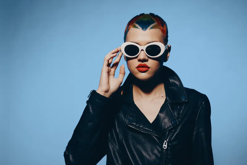 Woman with colorful short hair wearing white sunglasses and black leather jacket standing against plain blue background, showing bold style and contemporary fashion.