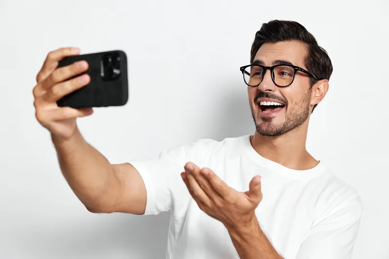 Smiling man wearing glasses holding smartphone and taking selfie, expressing joy while looking at the camera. Modern lifestyle and technology concept.