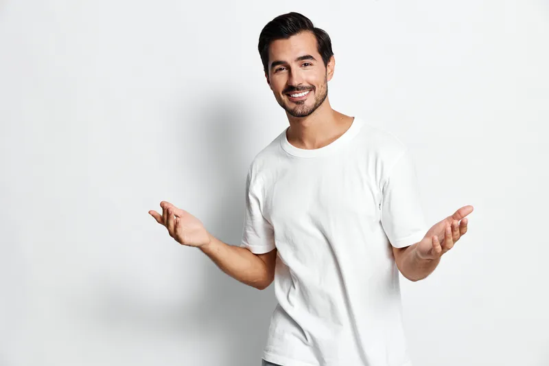 Friendly young man wearing a casual white t-shirt smiles broadly while gesturing with his hands, standing against a minimalistic light background. People lifestyle concept