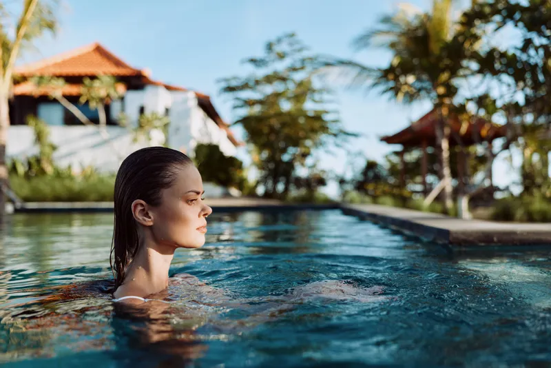 Young woman relaxes in outdoor swimming pool near tropical resort under clear sky, enjoying peaceful lifestyle and warm sunny weather during vacation time.