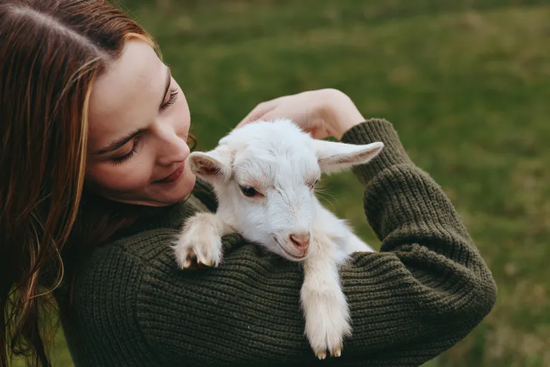 Cheerful woman outdoors holds a calm goat kid in a warm sunlit moment, relaxed pose, soft breeze, natural colors, rustic field background, intimate countryside lifestyle and friendly animal bond