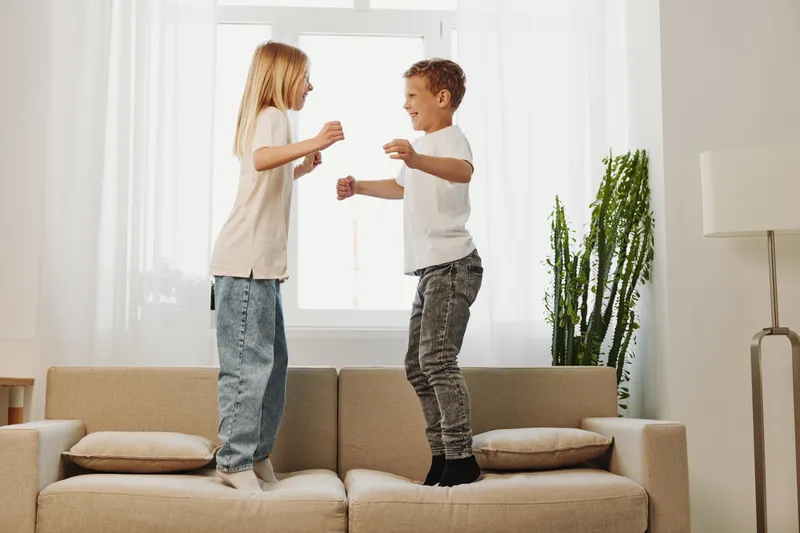 Two happy children jumping on beige sofa in bright living room, enjoying playful moment together. Concept for childhood, joy, fun, and family time.