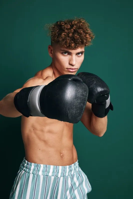 Young male boxer with curly hair wearing black gloves and striped shorts training in fighting stance against green background. Athletic, focused and shirtless athlete.