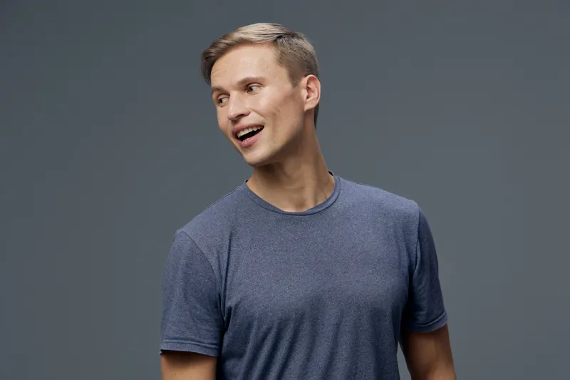 Friendly young man with short light brown hair wearing a casual blue t-shirt, smiling and looking to the side, standing against a plain gray background, studio portrait, lifestyle concept, positive em