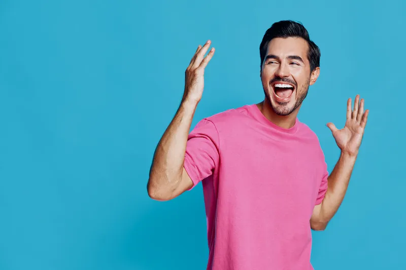 Happy cheerful young man wearing a bright pink t-shirt smiles broadly with raised hands, expressing joy in front of a solid blue background. Vivid lifestyle concept.