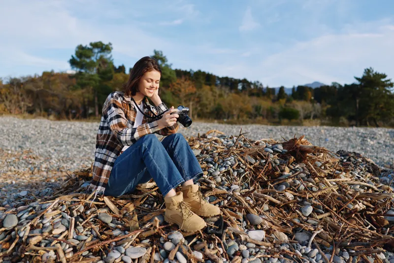 Photography of an outdoor woman sitting on a pebble beach, using a camera amid autumn trees, casual denim and boots, capturing calm coastal scenery and memories.