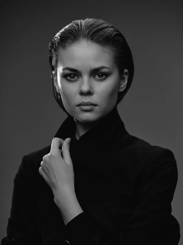 Black and white portrait of a serious woman wearing dark clothing with slicked-back hair in a studio setting.
