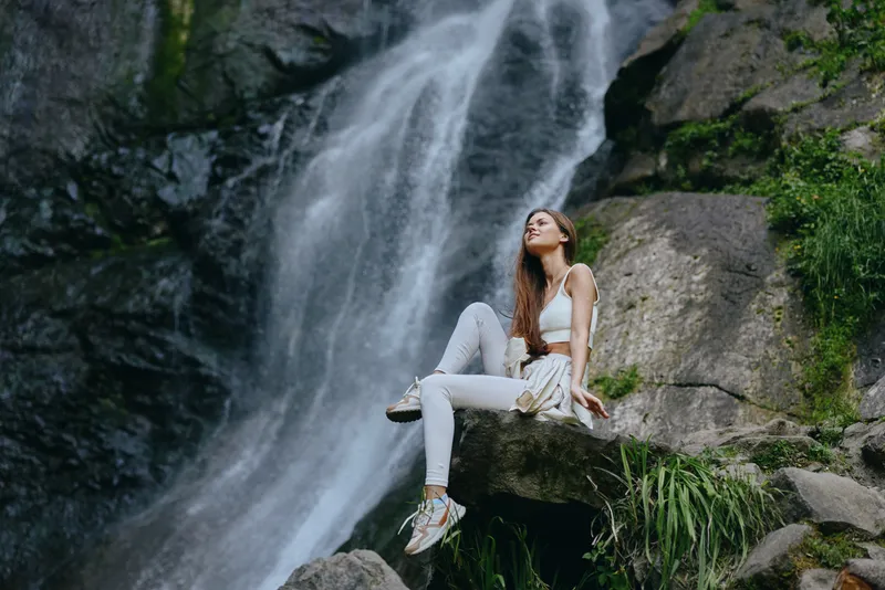 Young woman sitting on a rock near a waterfall, wearing casual white outfit, enjoying nature, peaceful outdoor moment, serene forest background.