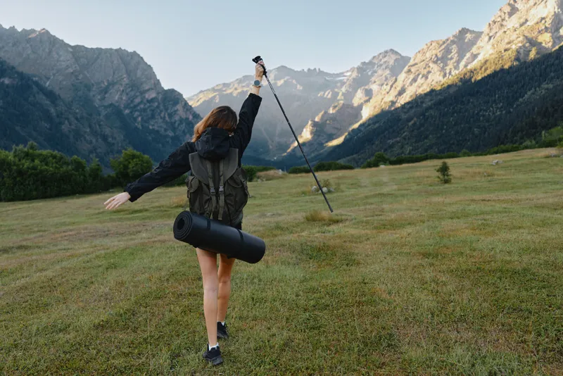 A lone hiker with a backpack is seen trekking across a grassy meadow toward rugged mountains. She raises a trekking pole aloft, creating a bold and adventurous outdoor atmosphere.