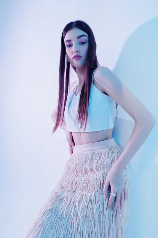 Young woman with long hair wearing a white crop top and textured skirt poses against a light background. Concept of woman beauty fashion and modern style.