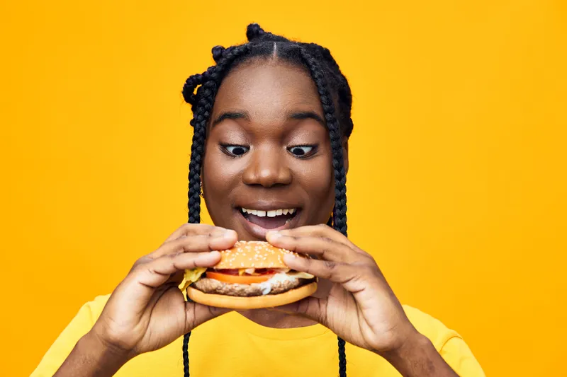 Happy young woman with braids enjoying a delicious burger against a bright yellow background, showcasing vibrant food photography and joyful emotions