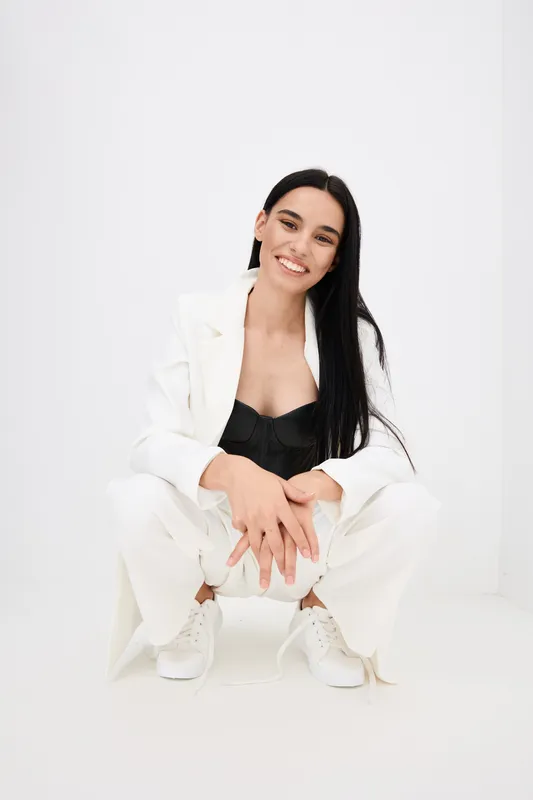 Smiling woman in white suit and black top squatting on white background with casual white sneakers, long dark hair, and cheerful expression showcasing modern fashion style.