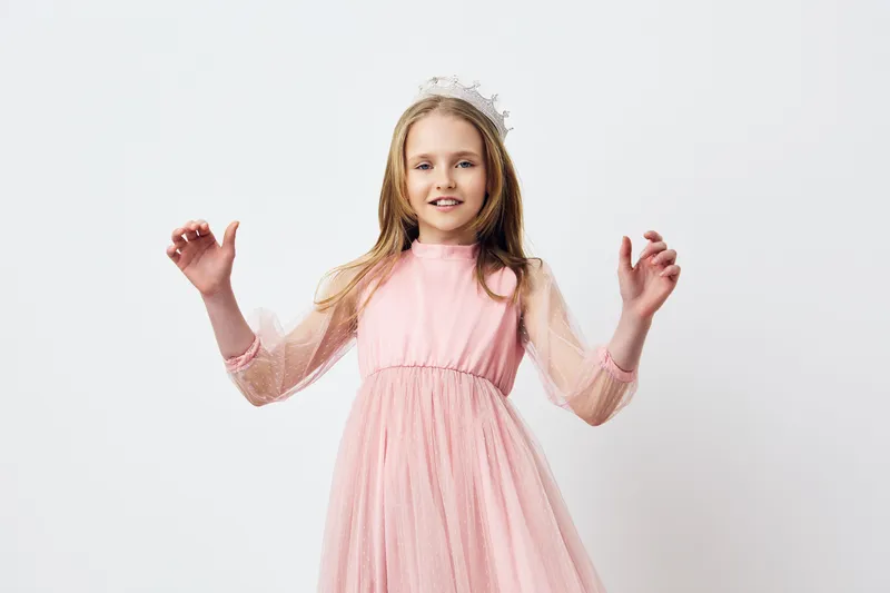A cheerful girl in a pink dress with sheer sleeves and a flower crown, joyfully posing against a soft white background, capturing the essence of childhood innocence and celebration