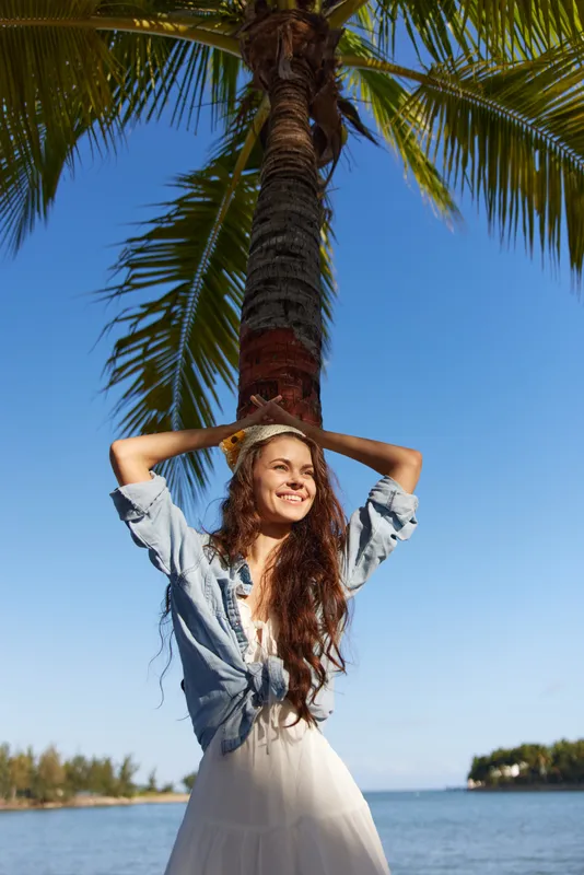 A joyful young woman stands at a tropical beach beneath a tall palm tree, wearing a light denim shirt and white dress, basking in bright sunshine and blue sky.