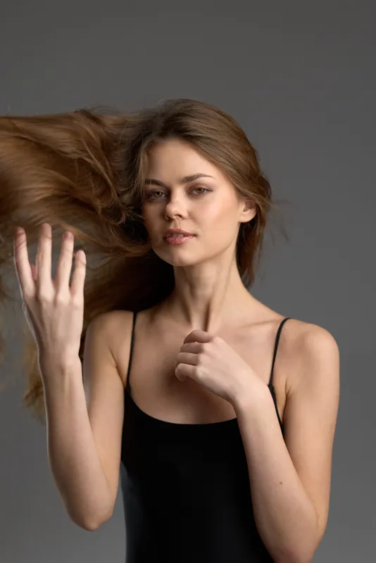 Young woman with flowing hair posing confidently with neutral expression wearing a black top against gray background.