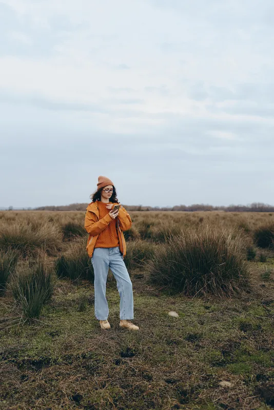 Woman lifestyle outdoors, standing in a grass field under overcast sky, wearing an orange jacket and denim jeans, relaxed pose, nature background, soft cloudy light