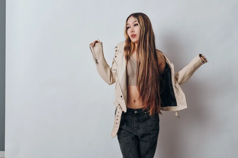Young woman with long braided hair wearing casual jacket and crop top posing with raised arms against plain light wall for fashion and style portrait.