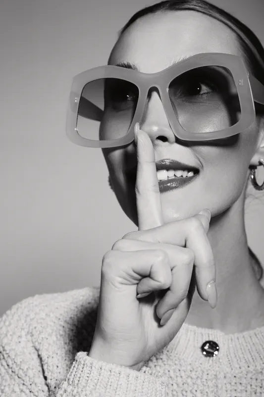 Stylish white woman with fair skin wearing large sunglasses and sweater holds finger to lips in gesture of silence.