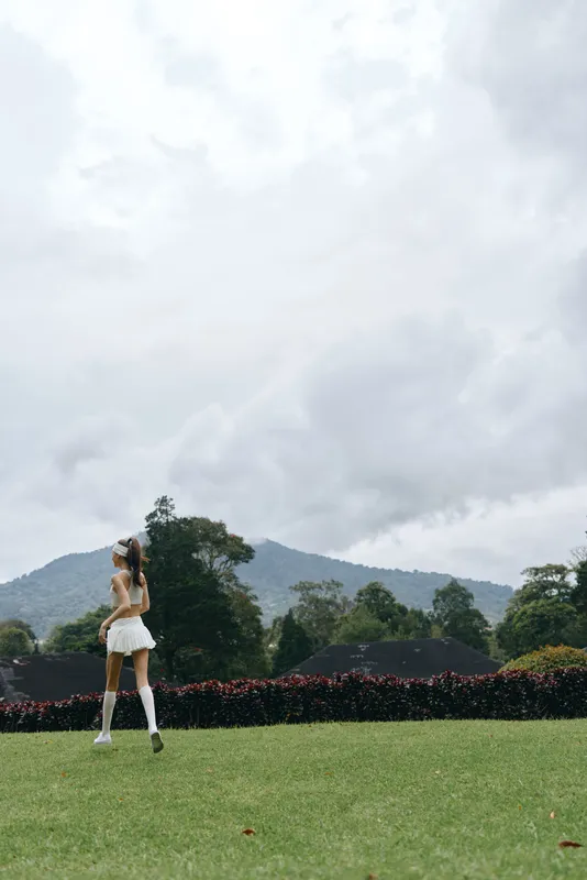 Young woman in white skirt and knee socks walking in lush green park under cloudy sky, evoking feelings of freedom and nature