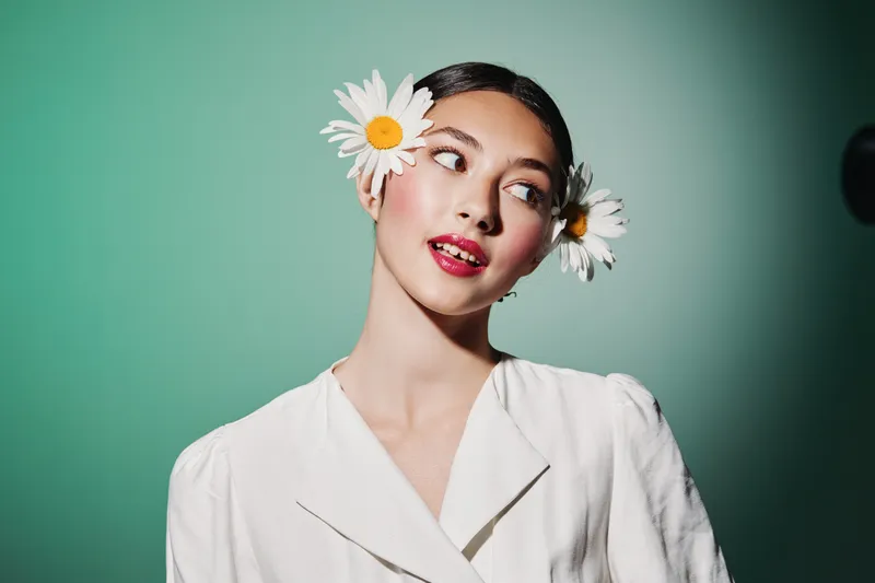 Young woman with daisy flowers in hair looks aside wearing white blouse. Clean natural makeup and stylish hair are highlighted on turquoise background with soft light. Beauty fashion creativity
