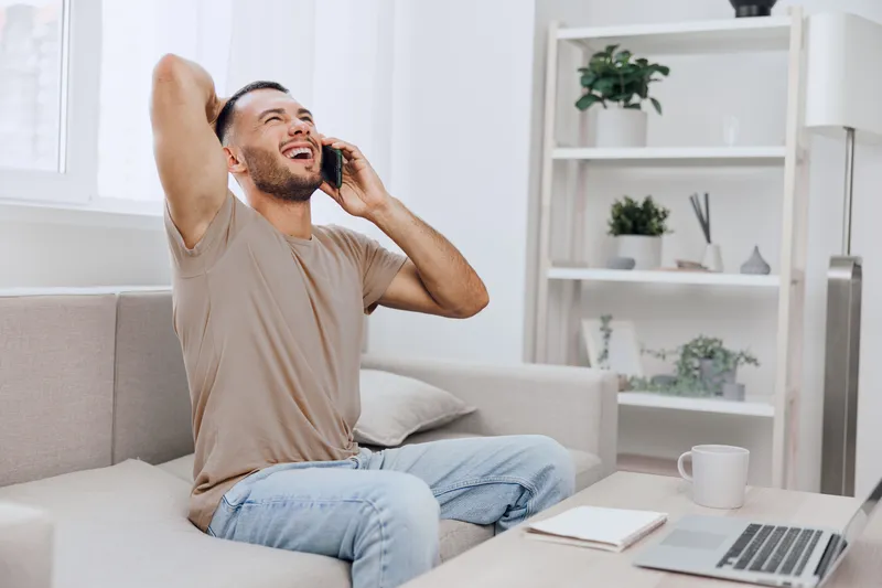Happy man talking on the phone in a modern living room, wearing casual clothes, enjoying a cheerful moment, surrounded by plants and natural light.