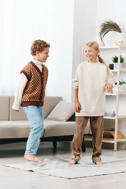 Two children dance playfully indoors, wearing warm sweaters and casual pants, on a light woven rug in a bright, cozy living room with soft natural light.