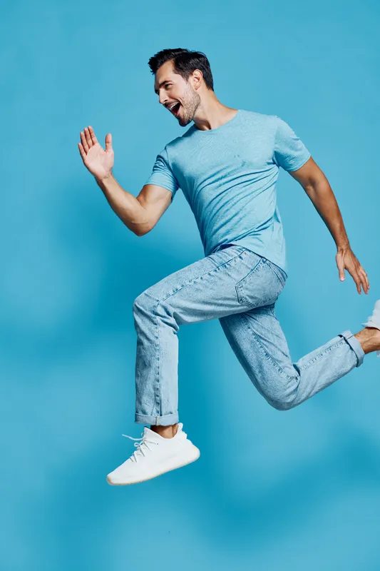 Full body joyful smiling young man wearing light blue t-shirt and jeans, jumping energetically against a bright blue background, expressing happiness and enthusiasm.