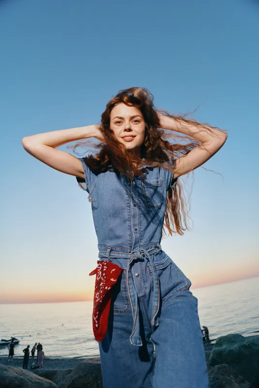 Lifestyle woman in modern boho-western denim street style posing outdoors by the sea with film color tones in natural sunset light, relaxed and confident mood.