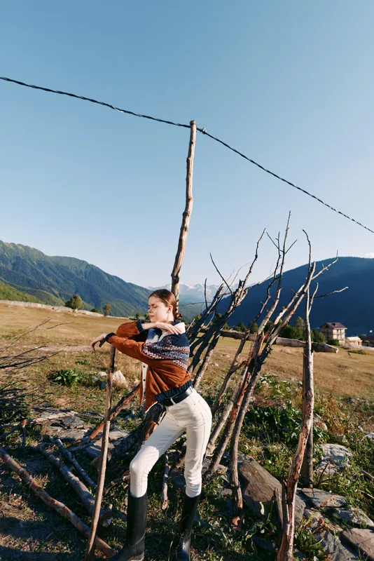 Woman in rural countryside leans on a rustic fence made of sticks, wearing boots and a sweater, framed by open field and distant mountains under a clear blue sky.