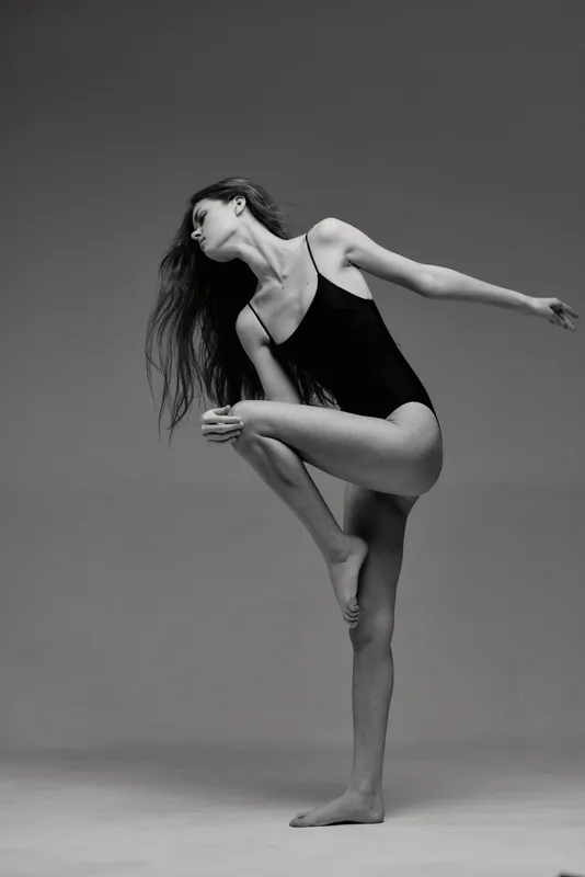 Black and white graceful woman in leotard posing with elegant balance and flowing hair against simple studio background.