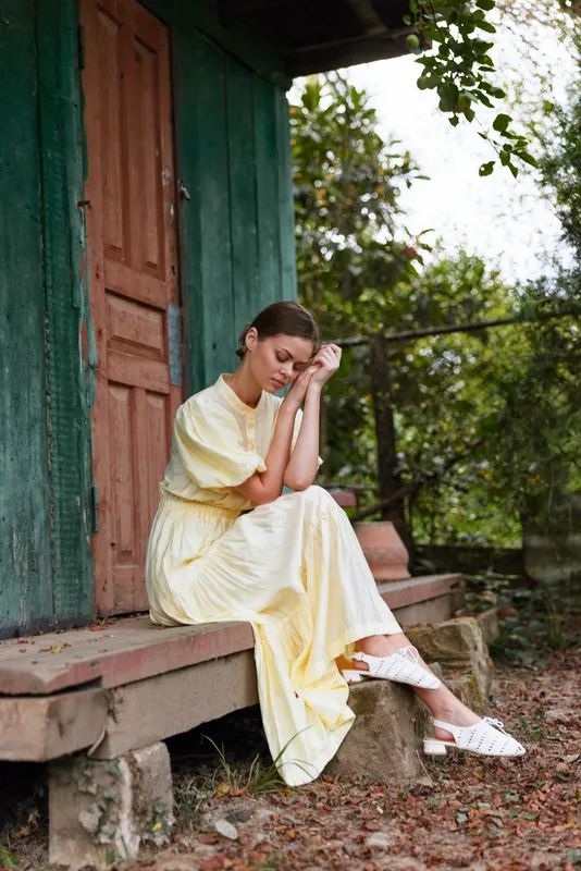 Woman in a yellow dress sits thoughtfully on outdoor steps beside a green door, creating a calm sunny mood and intimate portrait in a rustic setting.