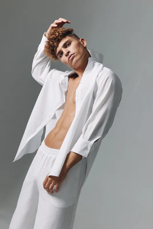 Young man with curly hair wearing white open shirt and pants posing with hand on head against gray background in studio. Fashion, casual style and confident expression in portrait.