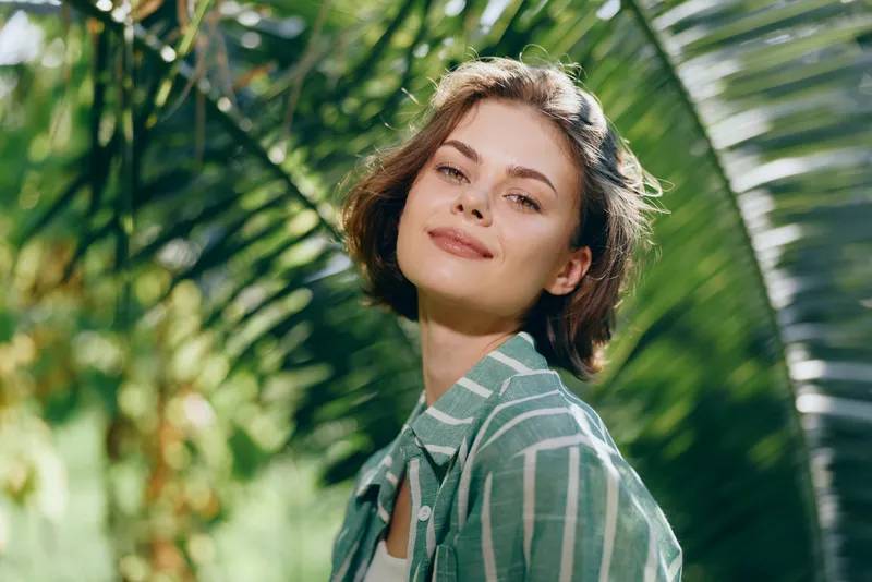 Portrait of a smiling woman outdoors, bathed in sunlight, framed by lush tropical leaves, conveying warmth, confidence, and natural beauty.