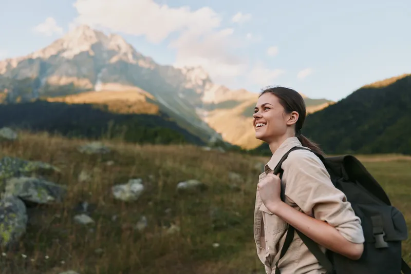 Woman hiking outdoors with a backpack, smiling as she walks through a sunlit meadow, with distant mountains and lush scenery, showcasing a relaxed lifestyle.