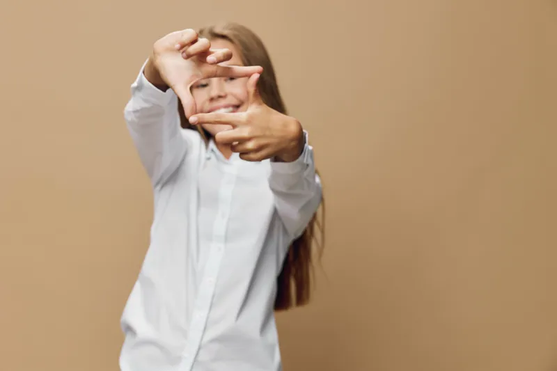 Girl with long hair and white shirt making frame gesture with hands in front of face on beige background with focus on playful expression and creativity.