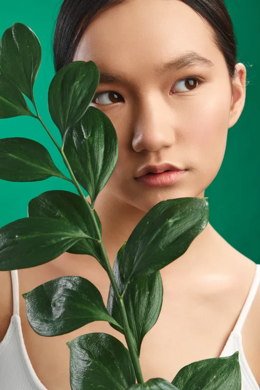 Asian woman gazes softly at the camera while holding a leafy plant in a studio with a vivid green backdrop, conveying freshness, calm and natural beauty.