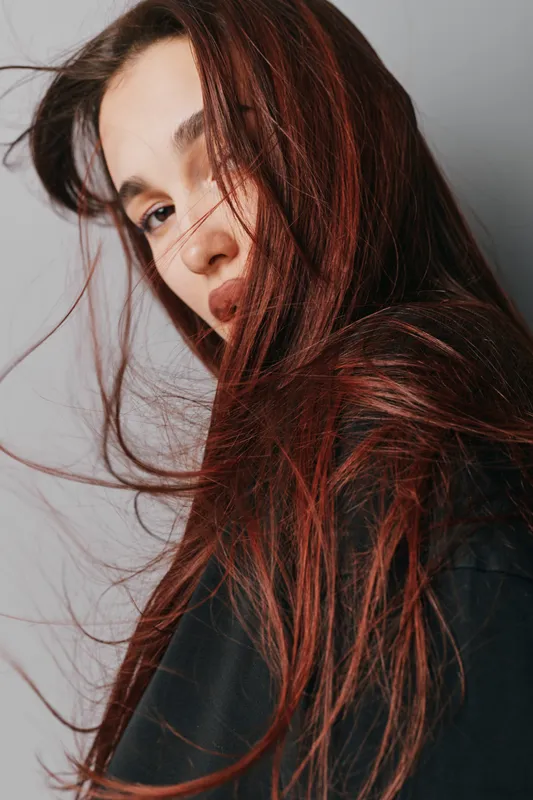 Portrait of young woman with long red hair looking at camera with serious expression. Closeup face and hair strands flowing, natural makeup and casual dark clothing on neutral background.