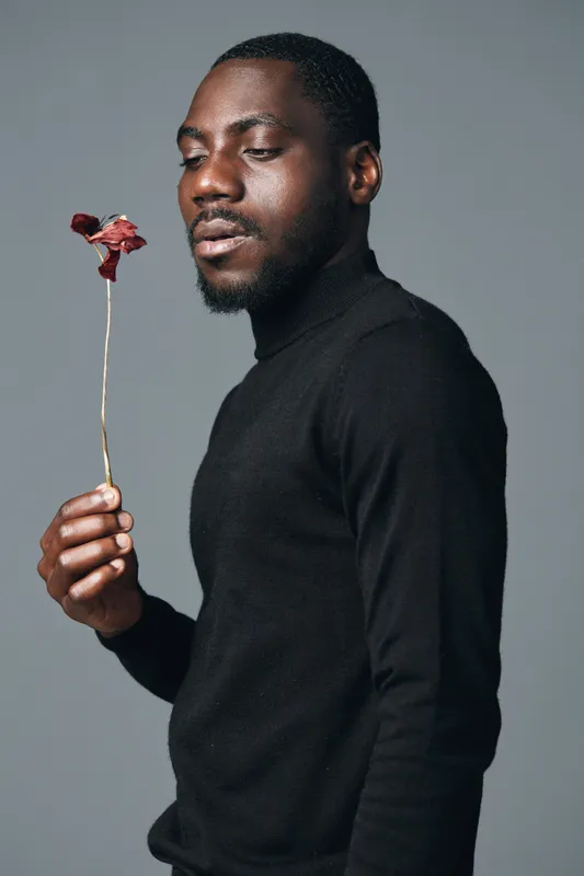 African man with dark skin holding dried red flower in hand wearing black turtleneck sweater against gray background in creative visual designer style portrait.