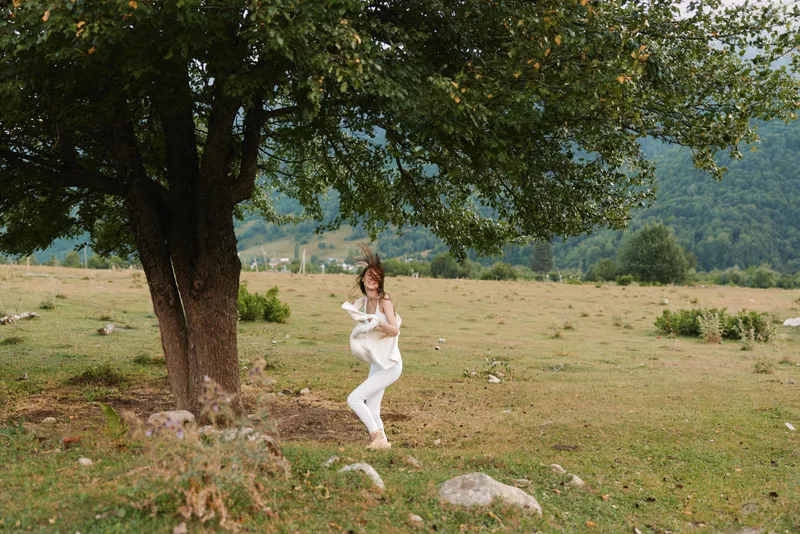 Casual woman in white stands by a tree in a sunlit meadow, capturing a serene lifestyle moment in nature, relaxed pose, peaceful mood, gentle breeze.